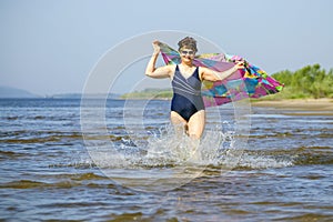 Athletic elderly woman runs along the waves on a summer sunny day against the background of mountains