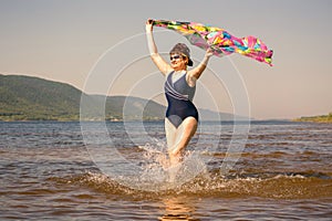 Athletic elderly woman runs along the waves on a summer sunny day against the background of mountains