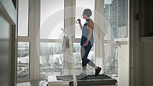 Athletic female is drinking water during exercising on treadmill. Physical exercises on smart trainer at home. Young woman is walk