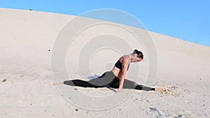 Athletic girl in black top and leggings performs stretching in twine pose, sits on the gymnastic twine on a deserted