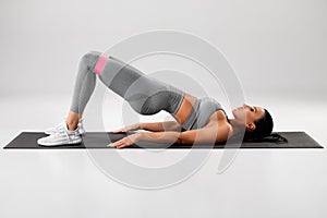 Athletic girl doing glute bridge exercise with resistance band on gray background. Fitness woman working out