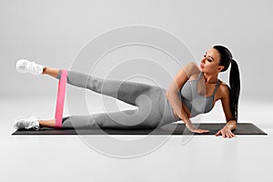 Athletic girl doing leg raises exercise for glute with resistance band on gray background. Fitness woman working out