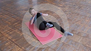 The athletic girl in a gray T-shirt and black tights lies on a pink rug and alternately bends her legs to strengthen her abdominal