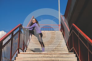 Athletic girl stands with her foot on the railing of the stairs, workout urban sport concept