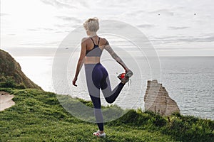 athletic girl in stylish sportswear stretching leg on cliff near sea Etretat France