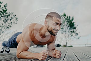 Athletic guy stands in plank pose to keep fit, leads healthy lifestyle and poses outdoor. Young bearded man in good physical shape