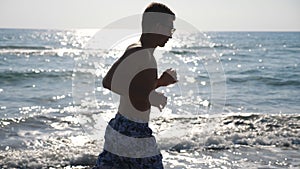 Athletic handsome guy jogging along sea coast at morning. Young sporty man running through ocean beach. Close up of male