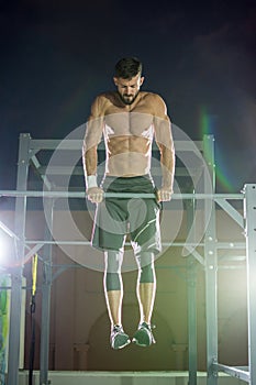 Athletic handsome man doing dip stand exercise on horizontal bar during calisthenics outdoors at night.