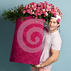 Smiling man delivery guy with rose at ear and lipstick kiss on his cheek is holding huge valentines day box with flowers