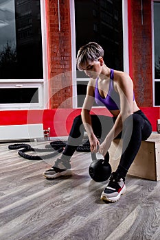 An athletic and happy young blonde female posing with atheltic training ropes and dumbbells in a gym.