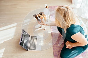 An athletic healthy mature 55s woman sits on a mat in lotus pose, doing breathing exercises, watching an online yoga