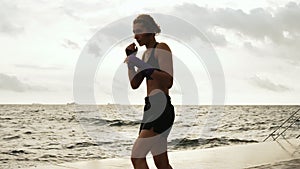 Athletic karate woman makes side kick of her legs against the sun by the sea in slow motion. Beautiful female boxer