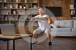 Athletic korean mature man doing deep squats during online workout at home, looking at laptop screen