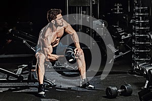 Athletic man bodybuilder execute exercise with dumbbells for biceps and sitting in dark gym