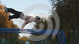 Athletic man doing gymnastics elements on bar in city park. Male sportsman performs strength exercises during workout
