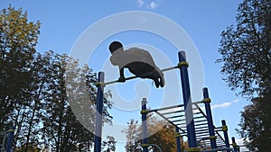 Athletic man doing gymnastics elements on horizontal bar in city park. Male sportsman performs strength exercises during