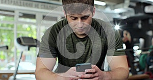 Athletic man relax sitting and texting message on smartphone at gym fitness. Serious male using mobile phone in sport