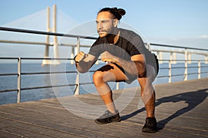 Athletic man in sportswear squatting at pier outside having workout