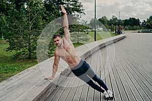 Athletic man stands in side plank pose, raises one arm, poses with shirtless torso, wears trousers and sneaks. Sportsman exercises