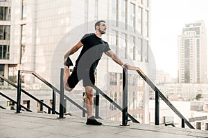 Athletic man stretching legs before workout outdoors. Runner in black sportswear excercising at morning holding handrail