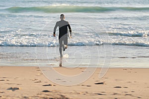 An athletic man surfer in a wetsuit running to the ocean holding a surfing board