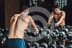 Athletic man waiting and looking at himself before lifting dumbbells in front of the mirror