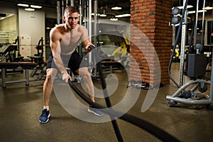 Athletic muscular man engaged with a thick rope with a bare torso in the gym.