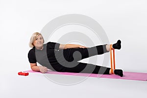 Athletic senior woman doing fitness workout at home with resistance rubber band lying on mat. Healthy lifestyle concept.