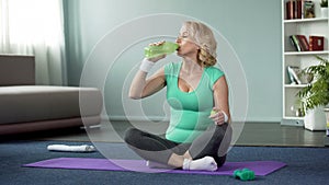 Athletic senior woman sitting on floor and drinking water after workout, health