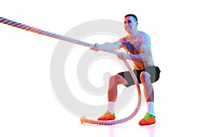 Athletic shirtless young man with strong body and hands pulling rope, training against white studio background in neon