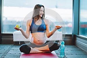Athletic smiling beauteous pregnant woman in sportswear. Doing yoga padmasana lotus exercise, choise green apple, donut