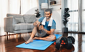 Athletic and sporty man drinking water on fitness mat at gaiety home.