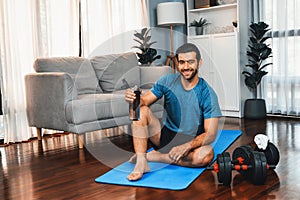 Athletic and sporty man drinking water on fitness mat at gaiety home.