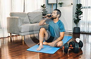 Athletic and sporty man drinking water on fitness mat at gaiety home.