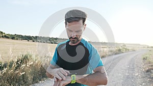 Athletic strong mature sportman running and looking at activity tracker on smart watch outside road through field.