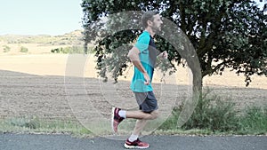 Athletic strong mature sportman wearing sport clothes while running outside road through field.