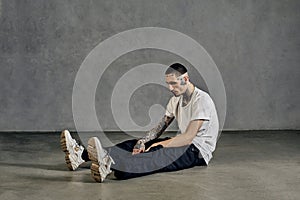 Athletic tattooed guy with beard. Dressed in white t-shirt, sneakers, black sports trousers. Sitting on floor, gray