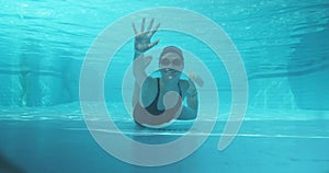 Athletic teenager girl swimmer underwater in blue outdoor pool looking at camera, waving his hand, hi hello gesture