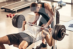 athletic trainer helping sportsman lifting barbell with heavy weight plates