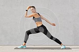 Athletic woman practices yoga, makes wide steps, shows good flexibility, poses against grey background, dressed in sportsclothes,