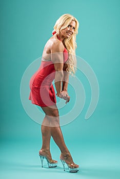 Athletic woman in red dress posing in studio. Attractive fitness lady with long blond hair showing her muscles.