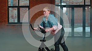 Athletic woman riding on spinning stationary bike training routine in haze gym, weight loss indoors