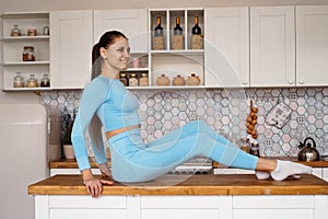 Athletic woman in sportswear posing on the table top of a light modern kitchen