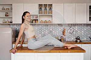 Athletic woman in sportswear posing on the table top of a light modern kitchen
