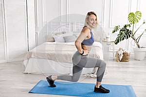 Athletic woman in stylish sportswear doing lunges exercises at home in bedroom. Attractive blonde-hair girl standing on