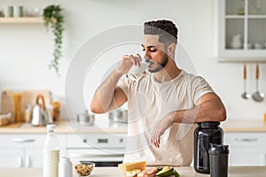 Athletic young Arab man drinking protein shake or milk, standing near table with healthy products at kitchen