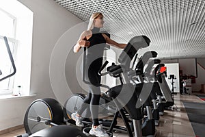 Athletic young beautiful woman in a black t-shirt in stylish leggings in white sneakers is training on a stepper simulator.