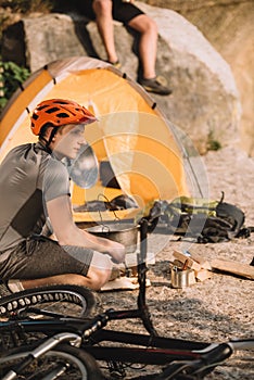 athletic young bike traveller sitting on rocks with camping tent and blurred friend sitting