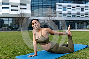 Athletic young black woman in sportswear doing fitness exercises on yoga mat, stretching her leg at city park