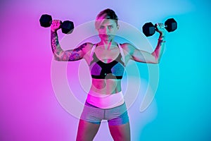 Athletic young blonde woman doing heavy dumbbell exercise for shoulders in the gym in neon lights.
